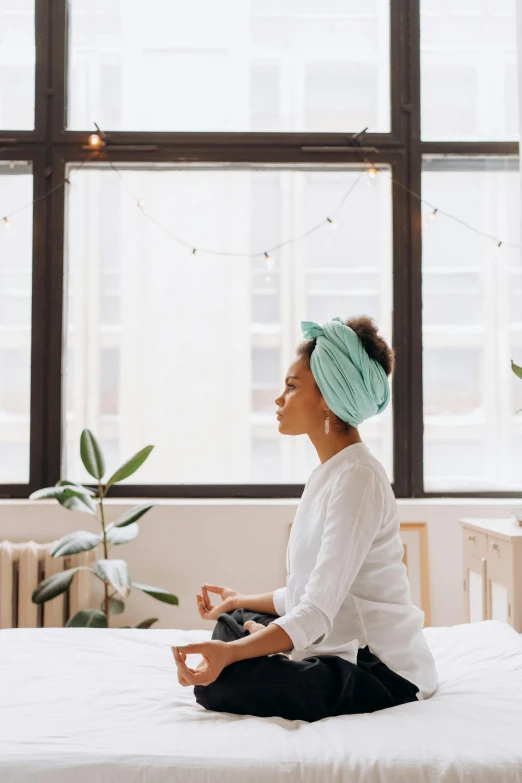 a woman sitting on a bed with a turban on her head, trending on unsplash, light and space, window sill with plants, meditating in lotus position, teal headband, on a desk