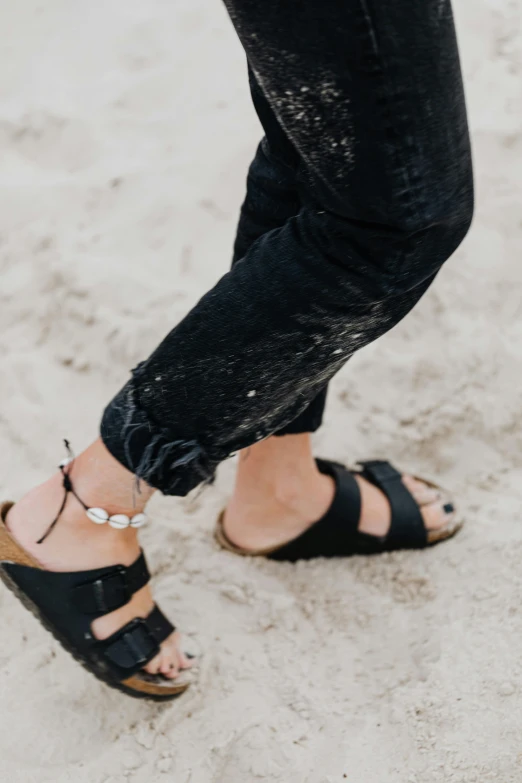 a woman standing on top of a sandy beach, by Nina Hamnett, trending on unsplash, birkenstock sandals, black pearls, black denim pants, dressed in a worn