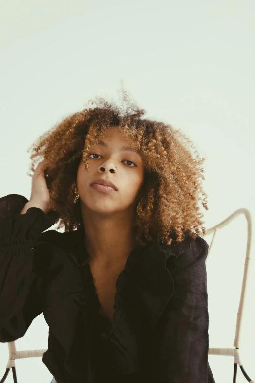 a woman with curly hair sitting on a chair, by Lily Delissa Joseph, trending on unsplash, renaissance, caramel. rugged, black teenage girl, natural muted tones, slightly minimal