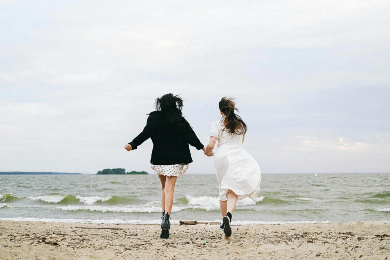 a couple of women standing on top of a sandy beach, by Lucia Peka, unsplash, romanticism, running fast towards the camera, lucy hale and maisie williams, wearing a cute white dress, celebrating an illegal marriage