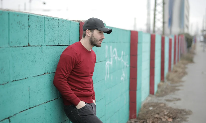 a man leaning against a green brick wall, a photo, red cap, background image, claudio bravo, red sweater and gray pants