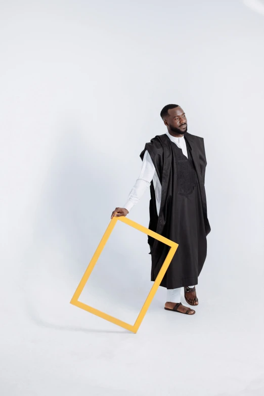 a man in a black robe holding a yellow frame, inspired by Ras Akyem, post graduate, full body photoshoot, wearing a grey robe, ( ( theatrical ) )