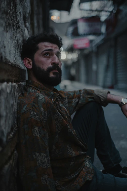 a man with a beard sitting against a wall, by Elsa Bleda, pexels contest winner, still from a music video, real life photo of a syrian man, mark edward fischbach, late evening