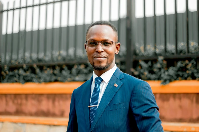 a man in a suit and tie standing in front of a fence, by Chinwe Chukwuogo-Roy, hurufiyya, man with glasses, parliament, guillaume tholly, professional profile photo