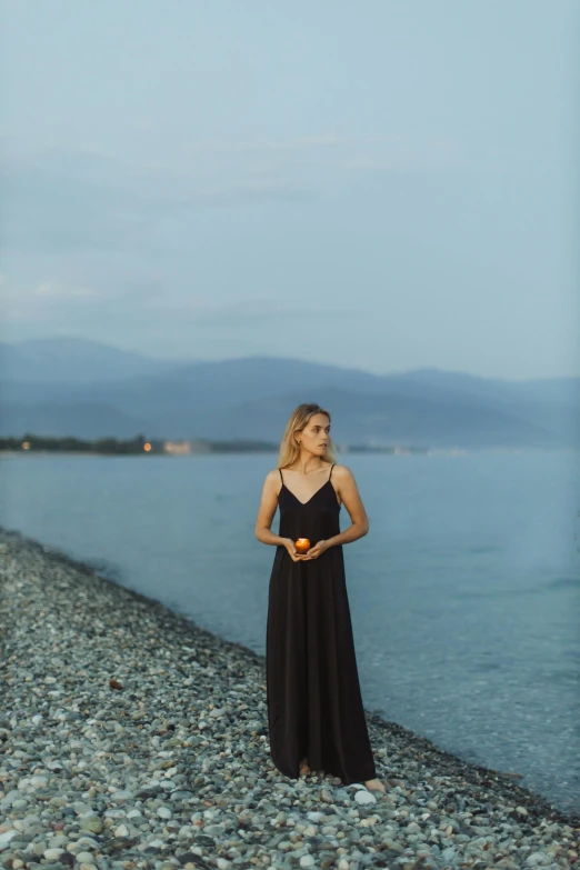 a woman standing on a beach next to a body of water, dark black long dress, greece, britt marling style, f / 2 0