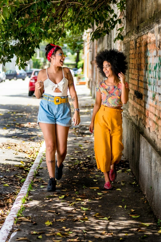 two women walking down a sidewalk next to a brick wall, a portrait, trending on pexels, brazilian, vintage clothing, croptop, with afro