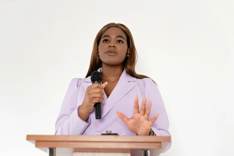 a woman standing at a podium with a microphone, pexels, emmanuel shiru, on a pale background, hands reaching for her, black young woman