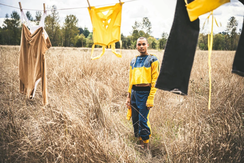 a man standing in a field of dry grass, a portrait, unsplash, visual art, with yellow cloths, laundry hanging, woman in streetwear, on display