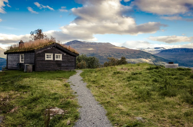 a small cabin sitting on top of a lush green hillside, by Jesper Knudsen, pexels contest winner, hurufiyya, nordic noire, rocky ground with a dirt path, with mountains as background, old house