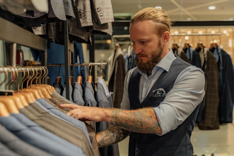 a man standing in front of a rack of clothes, inspired by Johan Lundbye, happening, dressed in blue, official store photo, wearing a vest, look at the details