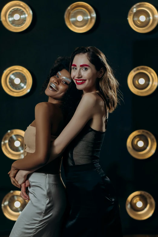 a couple of women standing next to each other, an album cover, trending on pexels, party lights, cheeky smile with red lips, at a fashion shoot, mix of ethnicities and genders