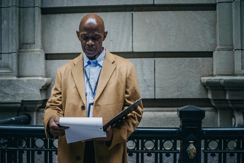 a man standing in front of a building holding a piece of paper, inspired by Gordon Parks, wearing a worn out brown suit, holding court, bald lines, humans of new york