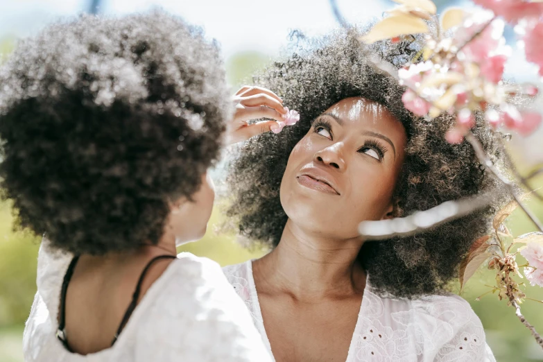 a woman brushing her hair in front of a mirror, pexels contest winner, afro made of flowers, sunny day time, woman holding another woman, looking upwards