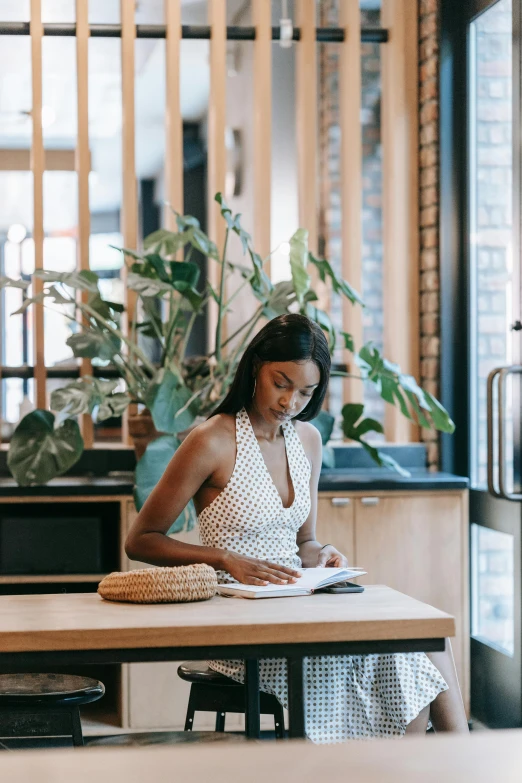 a woman sitting at a table reading a book, by Nicolette Macnamara, pexels contest winner, at the counter, black young woman, gif, standing elegantly