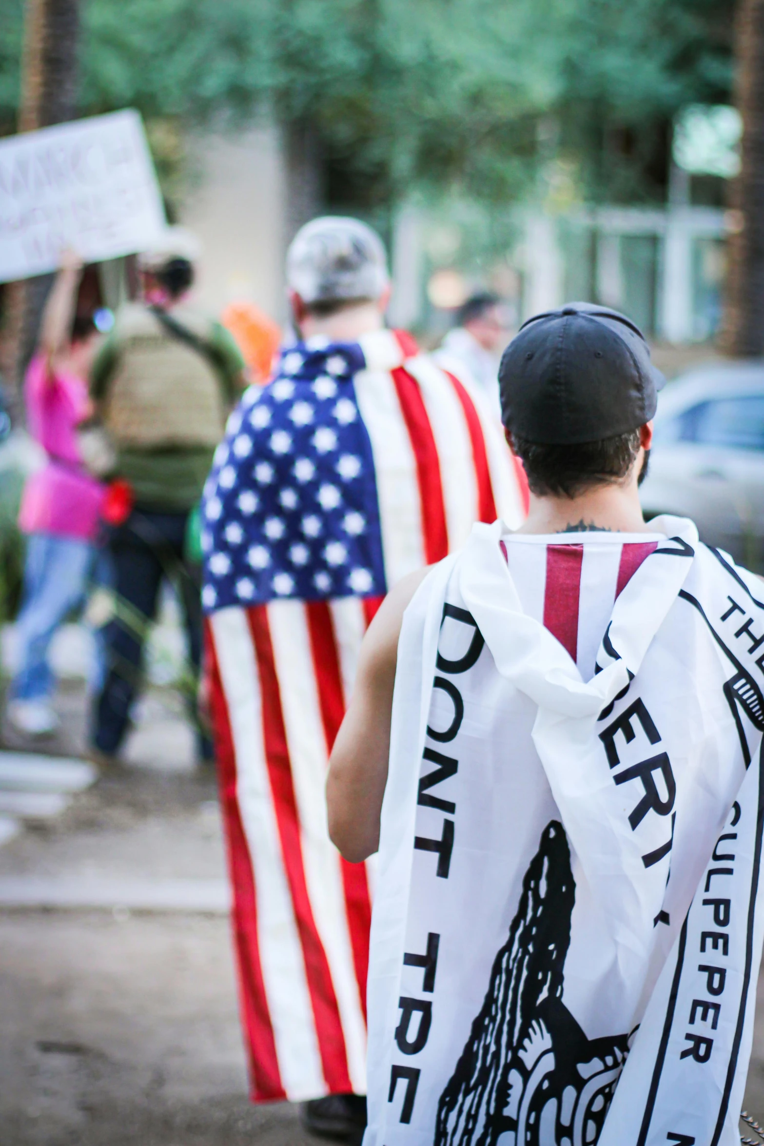 a group of people walking down a street holding flags, by Everett Warner, unsplash, photo of a man, wearing a tee shirt and combats, trans rights, his cape is the american flag