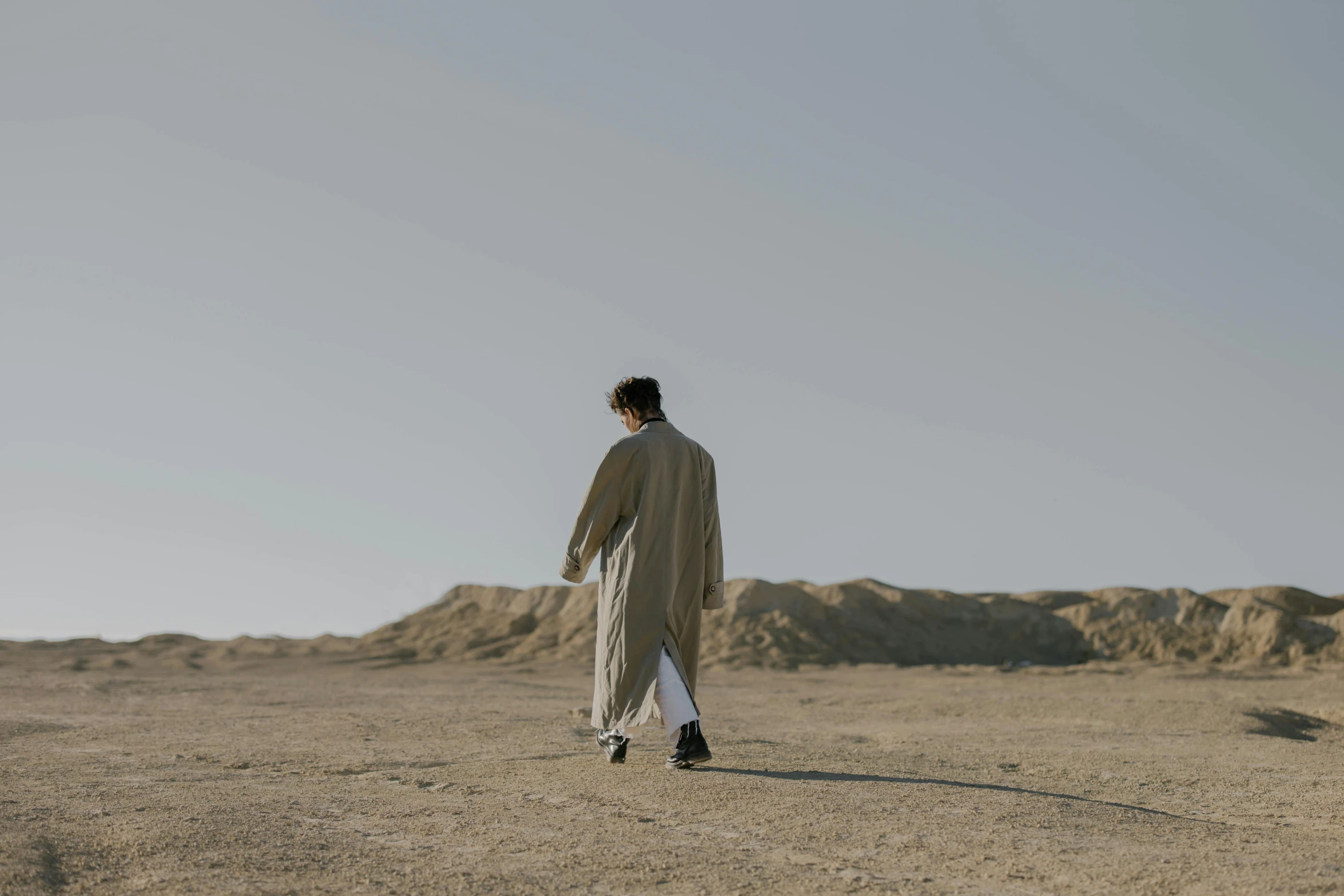 a man walking in the middle of a desert, an album cover, inspired by Amir Zand, unsplash, dirty linen robes, androgynous person, ignant, technological longcoat