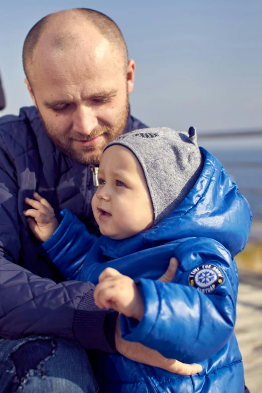 a man sitting on a bench holding a small child, inspired by Hallsteinn Sigurðsson, wearing blue jacket, model wears a puffer jacket, seaview, thumbnail