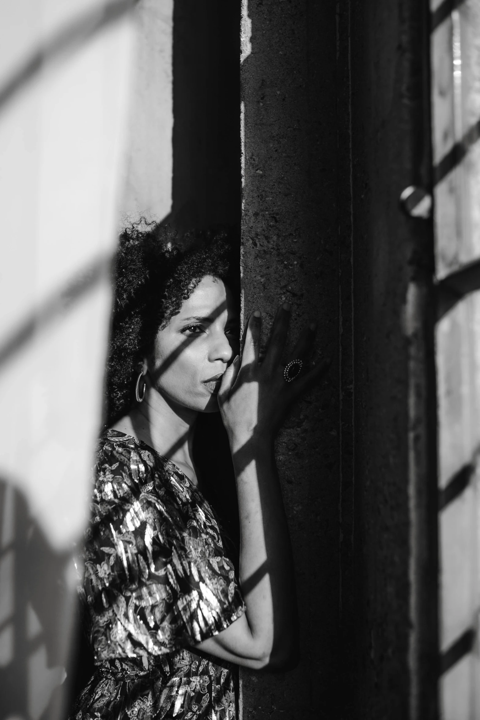a black and white photo of a woman leaning against a wall, pexels contest winner, aida muluneh, seen through a window, curly haired, harsh sunlight
