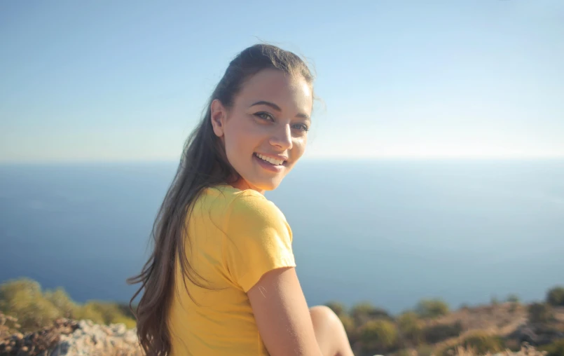 a woman sitting on top of a hill next to the ocean, greta thunberg smiling, profile image, sunny day time, yellow