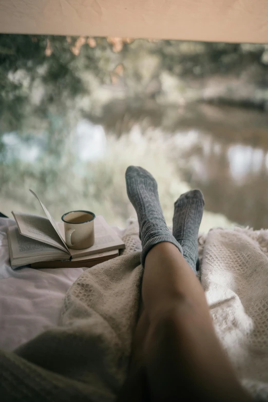 a person laying on a bed with a book and a cup of coffee, by Jessie Algie, unsplash contest winner, sitting near a river, wet feet in water, laying under a tree on a farm, misty weather