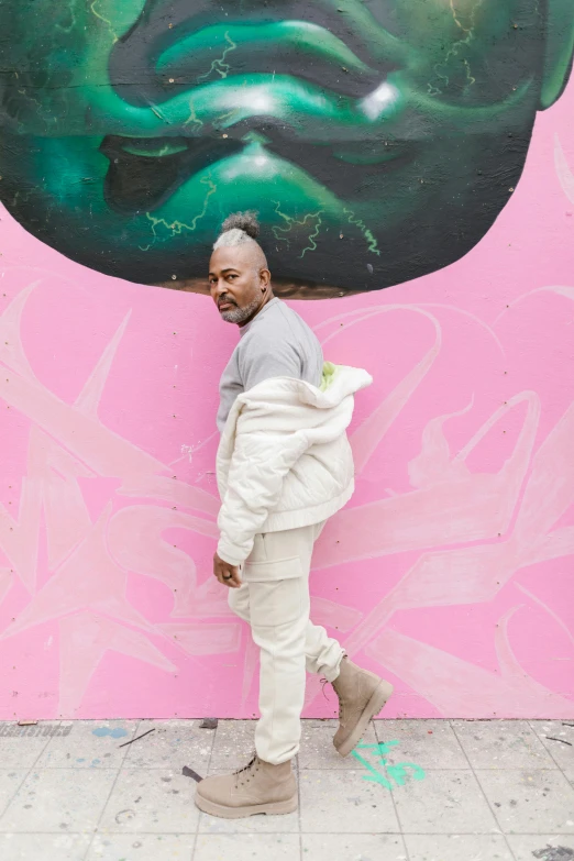 a man standing in front of a wall with a painting on it, an album cover, inspired by Michael Gustavius Payne, trending on unsplash, graffiti, pastel pink concrete, kanye, standing on a lotus, african aaron paul