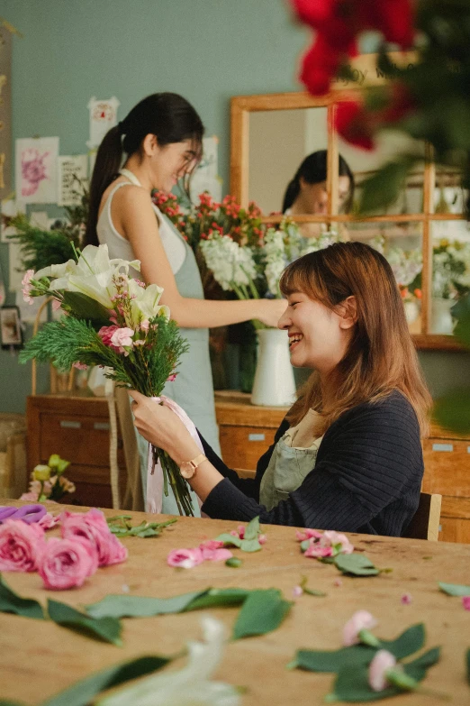 a woman sitting at a table with a bunch of flowers, in a workshop, smiling at each other, asian female, very aesthetically pleasing