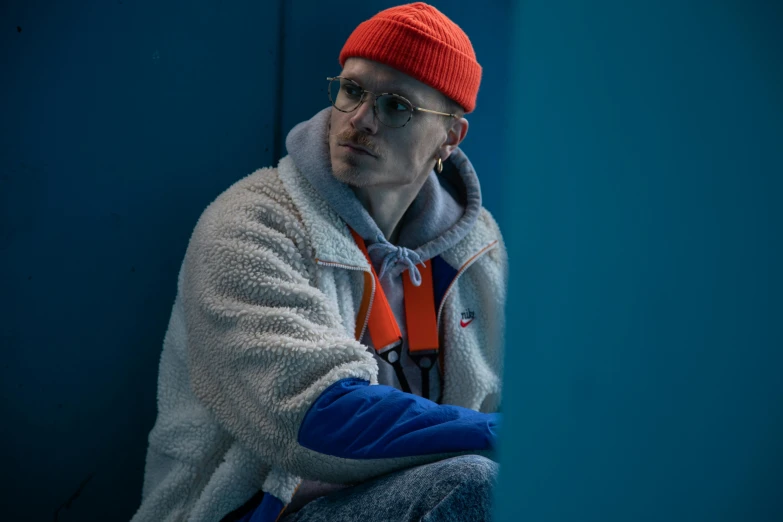 a man sitting on a bench wearing a red hat, a character portrait, inspired by Elsa Bleda, trending on pexels, die antwoord style wear, some orange and blue, with prison clothing, atmospheric cold lighting