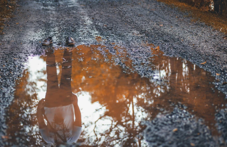 a person standing in a puddle on a road, inspired by Elsa Bleda, pexels contest winner, magic realism, gray and orange colours, water mirrored water, thumbnail, brown water