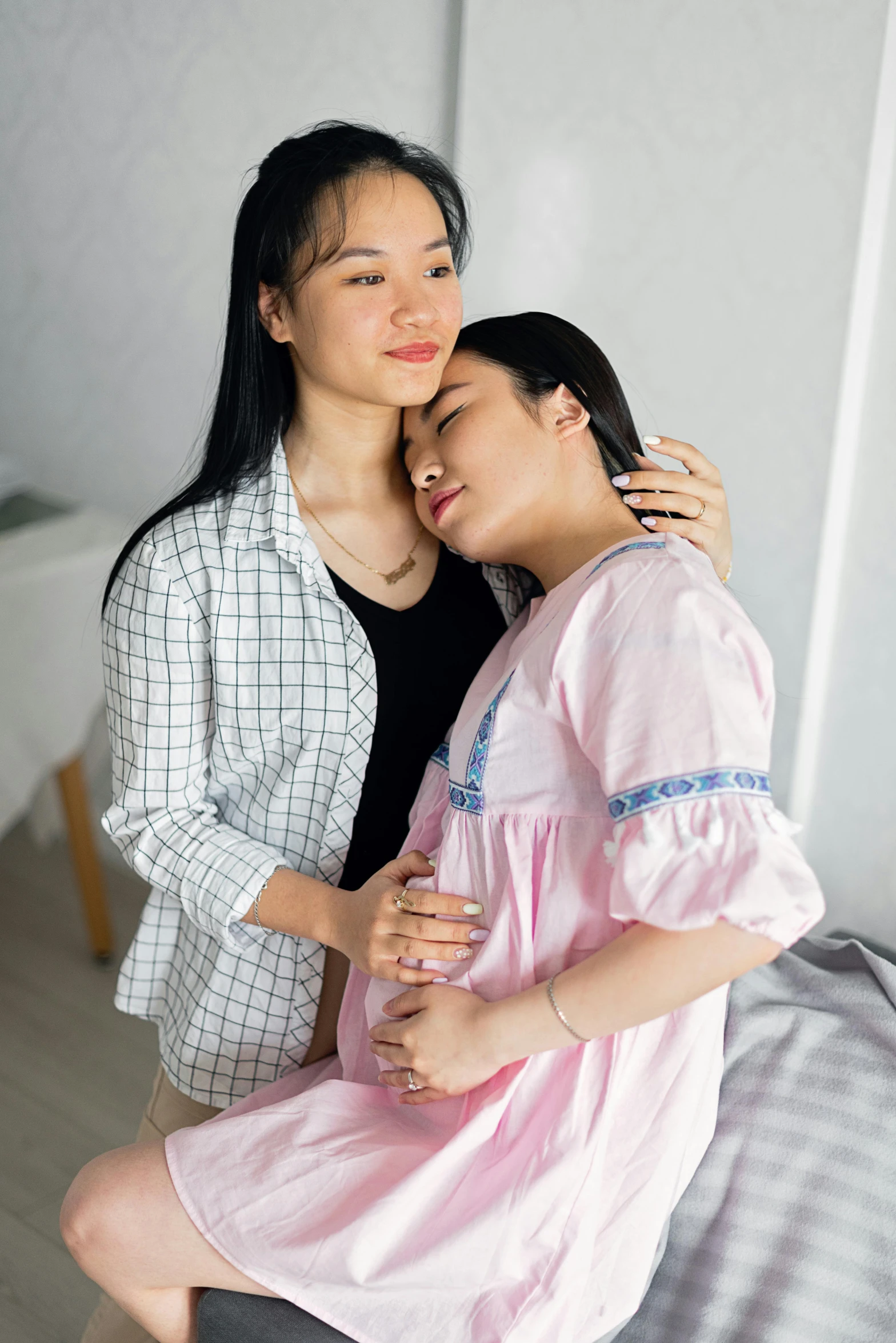 a woman sitting on top of a bed next to a woman, inspired by Ruth Jên, pexels contest winner, happening, pregnant belly, arm around her neck, asian, standing