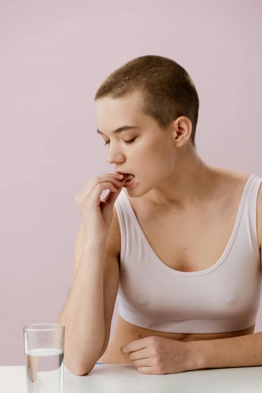 a woman sitting at a table with a glass of water, eating mars bar candy, very short slicked - back hair, wearing bra, nonbinary model