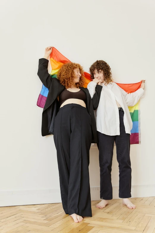 two women standing next to each other holding a rainbow flag, an album cover, by Harriet Zeitlin, unsplash, feminist art, large pants, black on white background, dressed with long fluent clothes, male and female