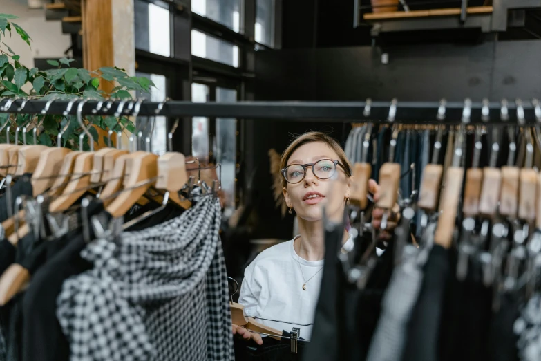 a woman standing in front of a rack of clothes, by Everett Warner, trending on pexels, happening, wearing square glasses, inspect in inventory image, sydney hanson, anna nikonova