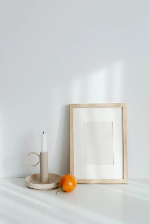 a picture frame sitting on top of a table next to an orange, a minimalist painting, trending on pexels, light and space, holding a candle holder, cream white background, with a wooden stuff, product display photograph