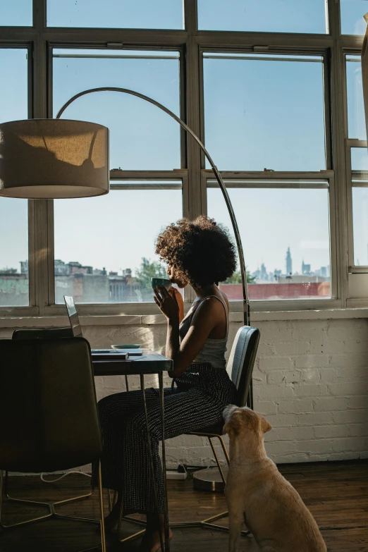 a woman sitting at a table with a dog in front of her, pexels contest winner, light and space, skyline showing from the windows, african american woman, picture of a loft in morning, table in front with a cup