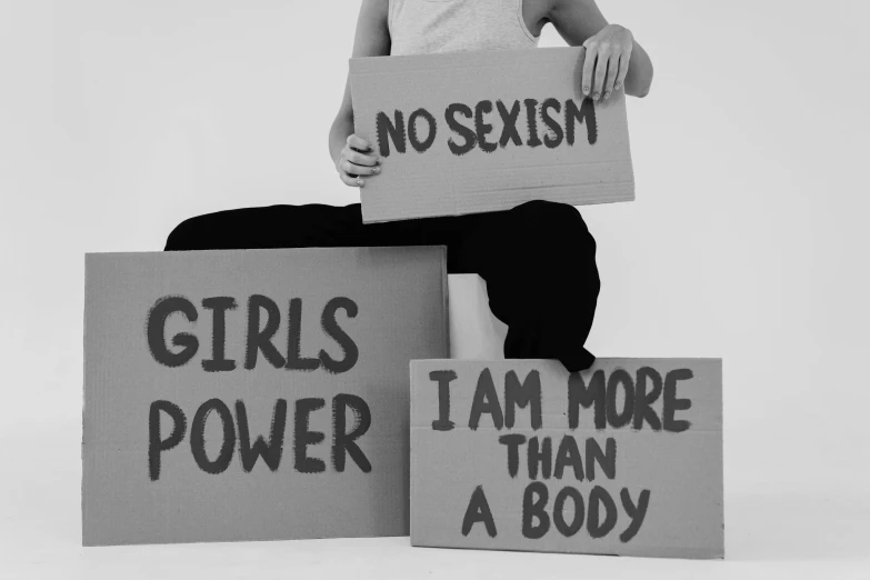 a woman holding a sign that says no sexism girls power i am more than a body, a poster, by Alexis Grimou, pexels, feminist art, upper and lower body, black - and - white photograph, no two bodies, brittney lee