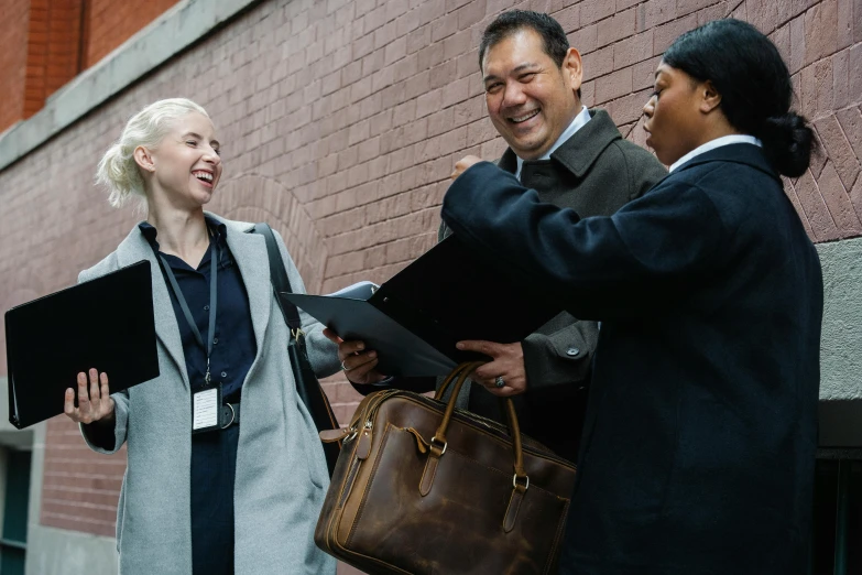 a group of people standing next to a brick wall, holding a briefcase, looking happy, amanda lilleston, medium close shot