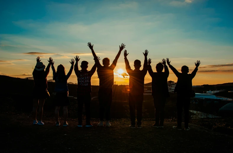 a group of people standing in front of a sunset, hands in the air, profile image, background image, ground level shot