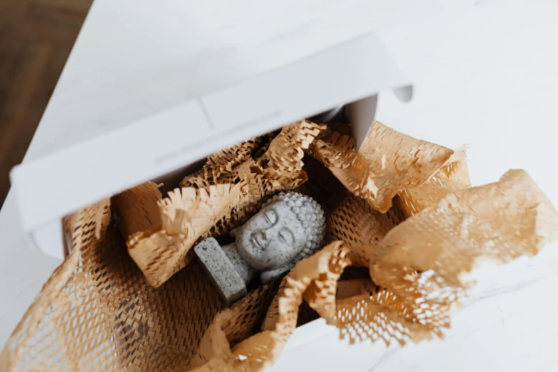 a small buddha statue sitting inside of a box, a marble sculpture, by Daniel Lieske, pexels contest winner, packaging design, dried flowers, brown paper, foil