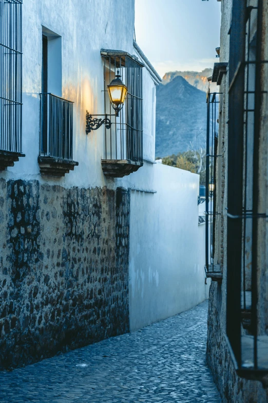 a narrow cobblestone street in an old town, inspired by Josep Rovira Soler, trending on unsplash, torches on the wall, mountains and a huge old city, white wall complex, square