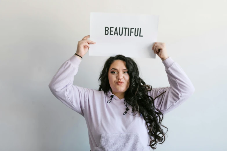 a woman holding a sign that says beautiful, by Olivia Peguero, trending on pexels, aestheticism, alluring plus sized model, looking upwards, on a pale background, beautiful - n 9