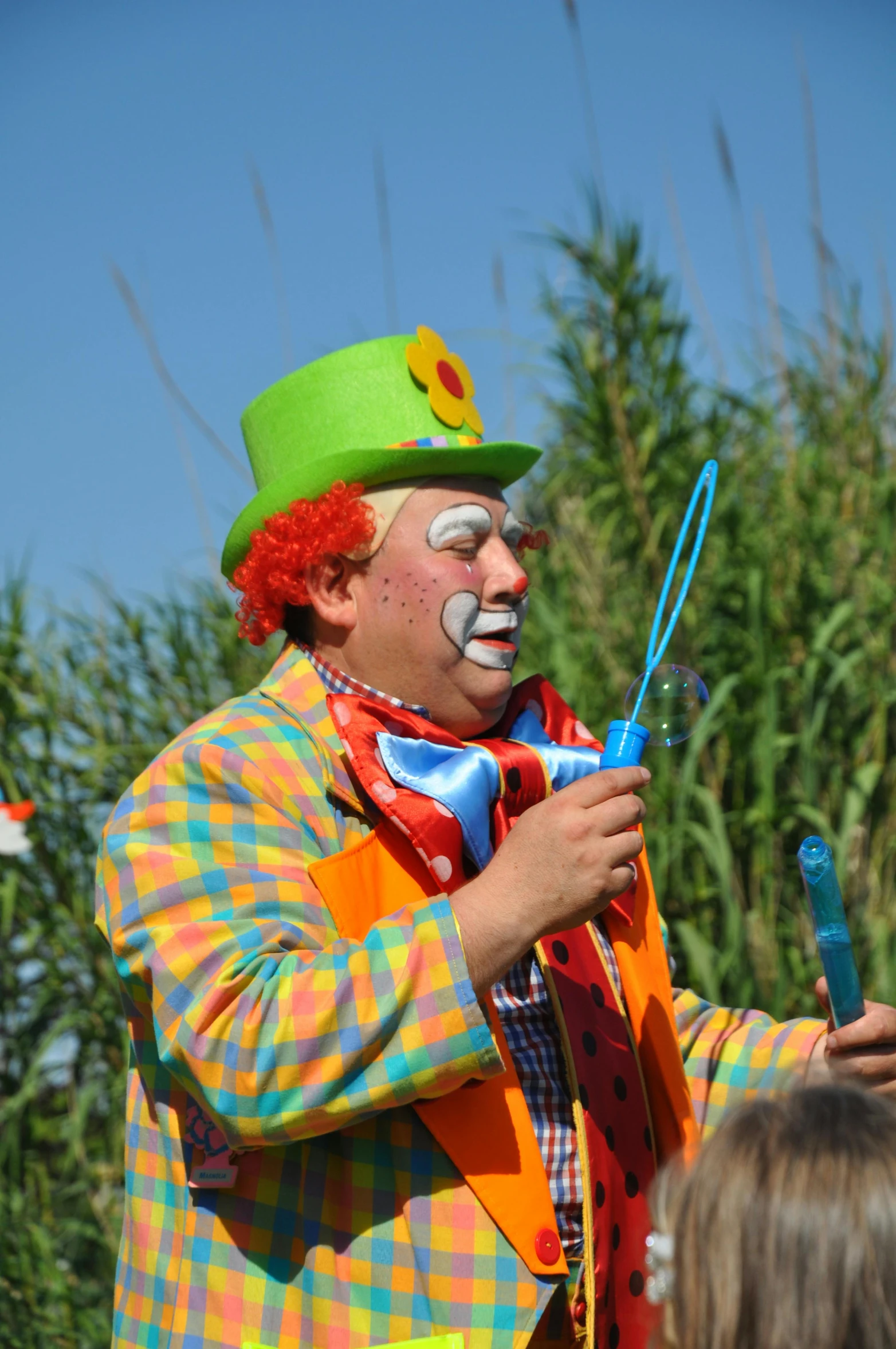 a clown standing in front of a group of people, with a straw, blue sky, long and orange mustache, performing