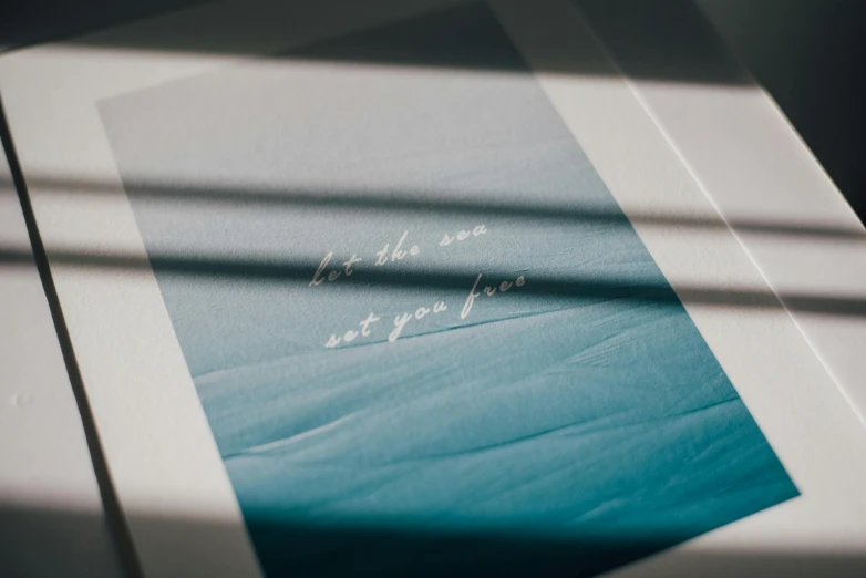 a book sitting on top of a table next to a window, a screenprint, unsplash contest winner, sots art, ride the wind and waves, soft blue texture, minimalist wallpaper, etched inscriptions
