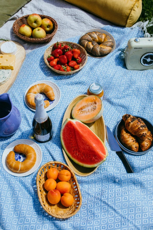 a blue and white blanket sitting on top of a grass covered field, plates of fruit, food, suns, bread