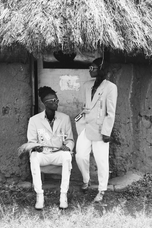 two people sitting on a bench in front of a hut, a black and white photo, afrofuturism, wearing white suit and glasses, dreads, brothers, playboi carti