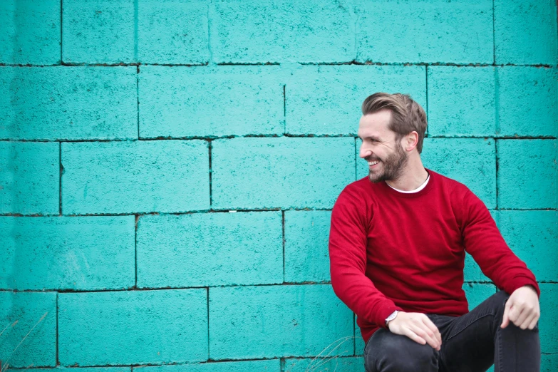 a man sitting on a skateboard in front of a blue wall, inspired by Alexander Robertson, happening, red sweater and gray pants, with teal clothes, profile image, smug smile