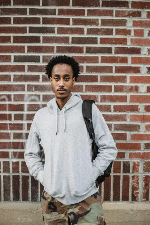 a young man standing in front of a brick wall, inspired by Afewerk Tekle, pexels contest winner, grey hoodie, ethiopian, at college, androgynous person