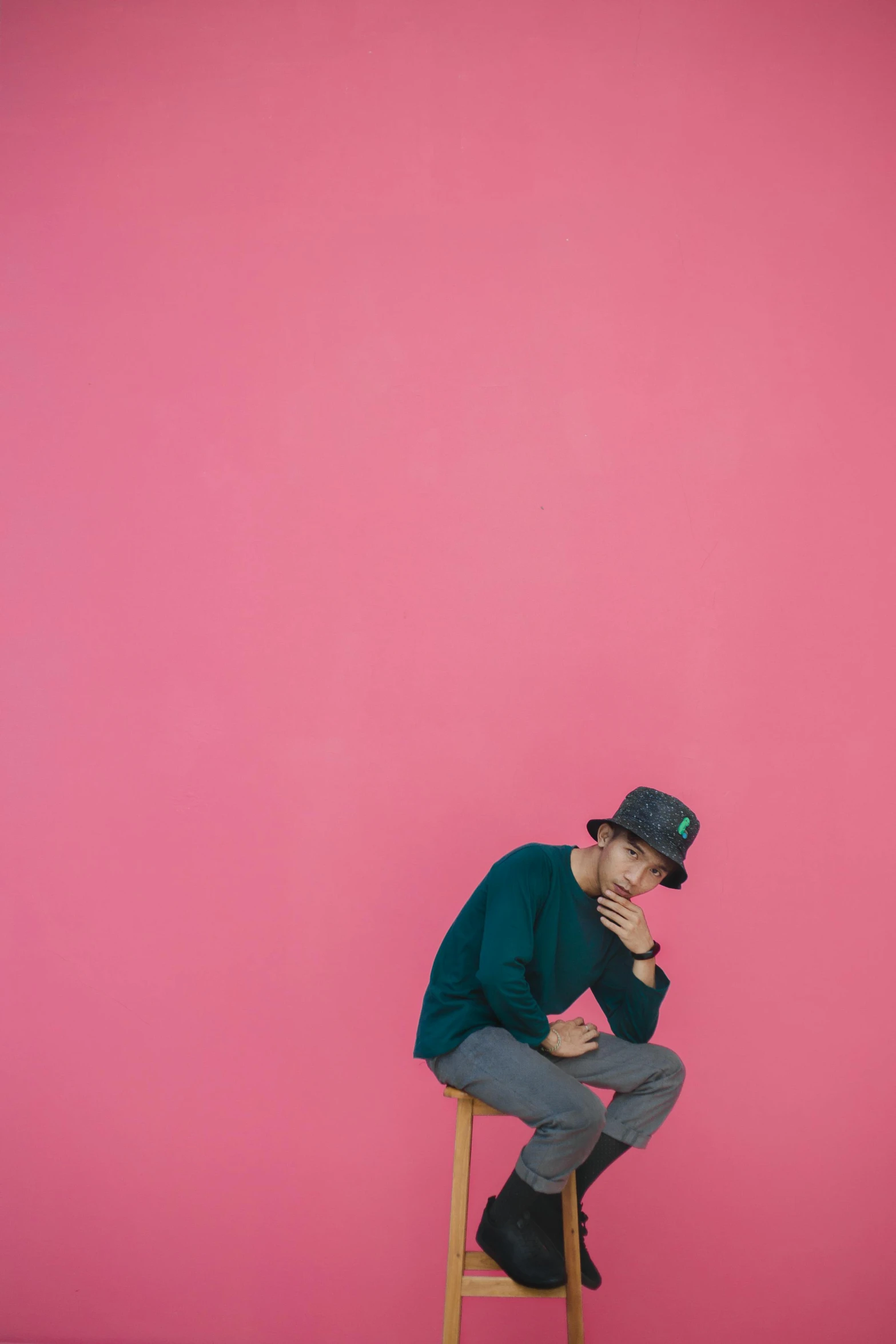 a man sitting on a stool in front of a pink wall, inspired by Junpei Satoh, trending on unsplash, minimalism, he is wearing a hat, thinker pose, full frame image, concert