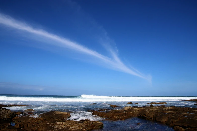 a large body of water sitting on top of a rocky beach, an album cover, unsplash, wispy clouds in a blue sky, australian beach, bulli, wave