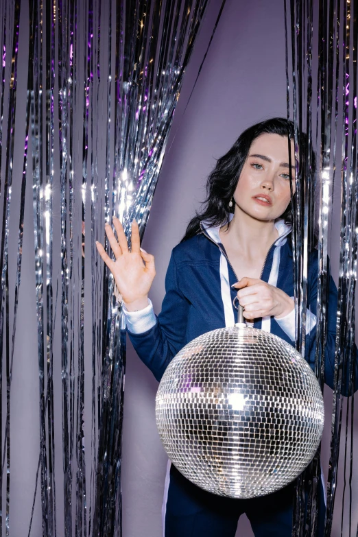 a woman holding a disco ball in front of a curtain, black haired girl wearing hoodie, ring lighting, meredit frampton style, on a pale background