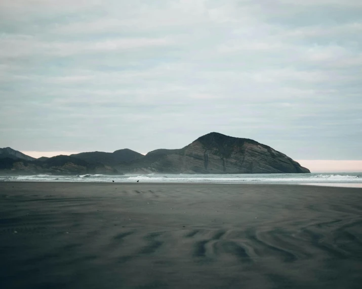 a man riding a surfboard on top of a sandy beach, by Jacob Burck, unsplash contest winner, minimalism, new zealand landscape, black volcano afar, in muted colors, in the distance is a rocky hill